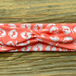 May include: Coral-colored headband with a repeating pattern of white baby footprints and syringes. The fabric is twisted at the center, creating a stylish accessory. This headband is perfect for expecting parents or those in the medical field.