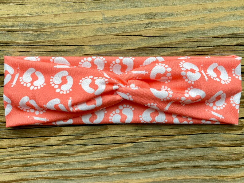 May include: Coral-colored headband with a repeating pattern of white baby footprints and syringes. The fabric is twisted at the center, creating a stylish accessory. This headband is perfect for expecting parents or those in the medical field.