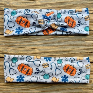 May include: Two gray headbands with a medical theme. The fabric is patterned with orange first aid kits, blue stethoscopes, teal face masks, and blue medical symbols. The top headband is twisted at the center.