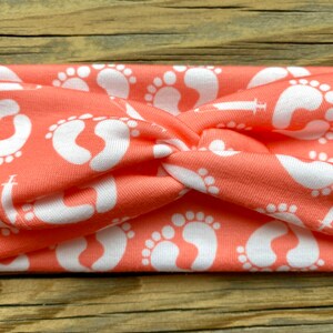 May include: Coral-colored headband featuring a repeating pattern of white baby footprints. The fabric is twisted at the center, creating a stylish accessory. This headband is a comfortable and fashionable choice for everyday wear.