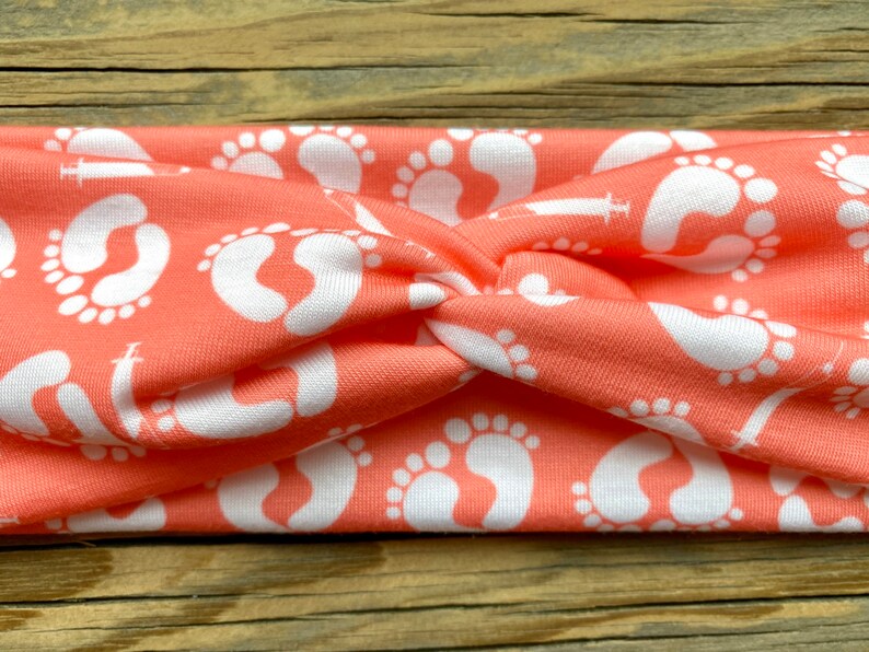May include: Coral-colored headband featuring a repeating pattern of white baby footprints. The fabric is twisted at the center, creating a stylish accessory. This headband is a comfortable and fashionable choice for everyday wear.