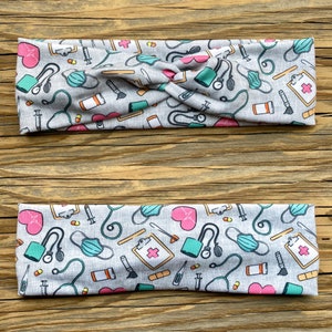 May include: A gray headband with a twisted design features a repeating pattern of medical symbols, including hearts, stethoscopes, bandages, pills, and syringes. The headband is made of a soft, stretchy fabric.