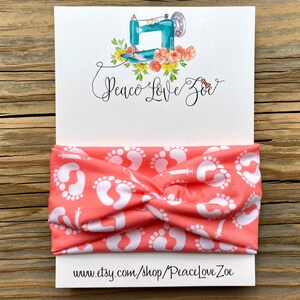 May include: Coral-colored headband with a white baby footprint pattern. The headband is displayed on a white card with the text "Peace Love Zoe" and a watercolor illustration of a sewing machine and flowers. The website address is also visible.