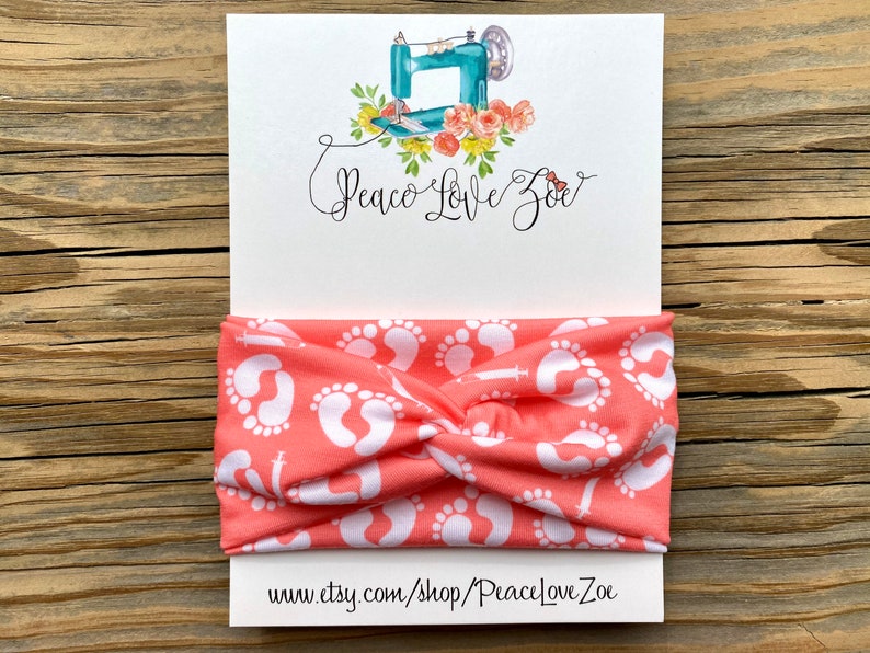 May include: Coral-colored headband with a white baby footprint pattern. The headband is displayed on a white card with the text "Peace Love Zoe" and a watercolor illustration of a sewing machine and flowers. The website address is also visible.