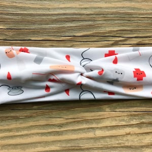 May include: White headband with a repeating pattern of medical-themed illustrations. The design includes red crosses, stethoscopes, bandages, and blood drops, all with smiling faces. The headband is twisted at the center.