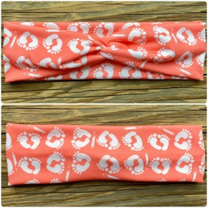 May include: Coral-colored headband with a white pattern of baby footprints and syringes. The headband is twisted at the top. The fabric is soft and stretchy, suitable for various head sizes. The design is repeated across the fabric.