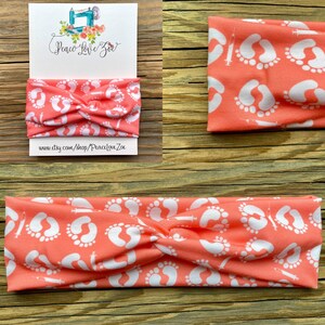 May include: Coral-colored headband with a white pattern of baby footprints and syringes. The headband is twisted at the top. A card with the text "Peace Love Zoe" and a website address is visible.