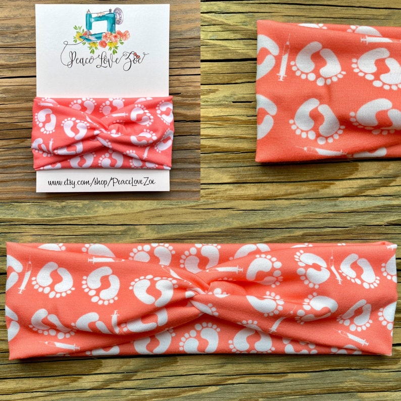 May include: Coral-colored headband with a white pattern of baby footprints and syringes. The headband is twisted at the top. A card with the text "Peace Love Zoe" and a website address is visible.
