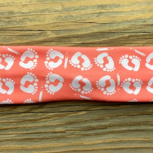 May include: Coral-colored headband featuring a repeating pattern of white baby footprints and syringes. The headband is made of a soft, stretchy fabric, perfect for everyday wear. The design is printed on a solid coral background.