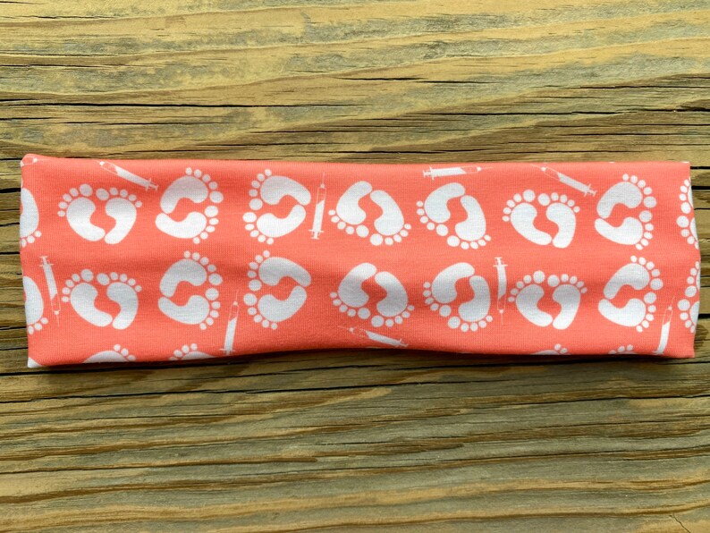 May include: Coral-colored headband featuring a repeating pattern of white baby footprints and syringes. The headband is made of a soft, stretchy fabric, perfect for everyday wear. The design is printed on a solid coral background.