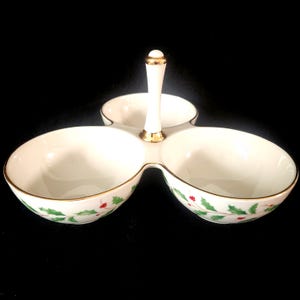 May include: A white ceramic serving dish with three sections, featuring a gold rim and a handle. The dish is decorated with green holly leaves and red berries, perfect for serving snacks or dips during the holidays.