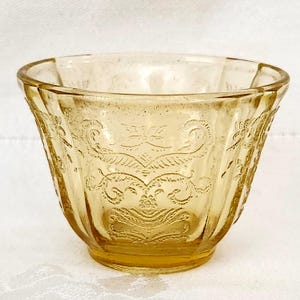 May include: A vintage amber-colored glass bowl with a decorative etched pattern. The bowl has a flared shape with a scalloped rim and a detailed floral and scroll design. The glass has a slightly textured appearance.