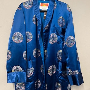 Op de afbeelding: Een blauwe satijnen pyjamajas met een herhalend cirkelvormig patroon in roze, wit en groen. De pyjamajas heeft een reverskraag, twee voorzakken en een label met tekst. De mouwen hebben manchetten.