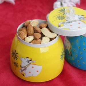 May include: Yellow ceramic jar filled with mixed nuts, with a lid featuring a cow illustration. A second blue jar with a cow illustration is visible. The jars are on a red surface.
