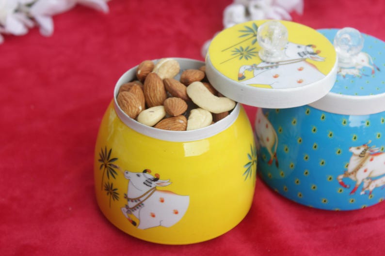 May include: Yellow ceramic jar filled with mixed nuts, with a lid featuring a cow illustration. A second blue jar with a cow illustration is visible. The jars are on a red surface.