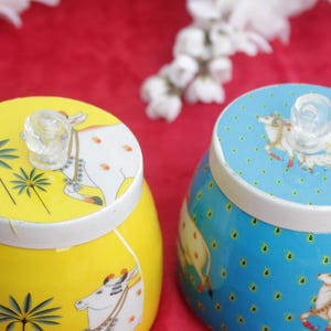 May include: Two decorative ceramic jars, one yellow and one blue, with white rims and clear glass knob handles. The yellow jar features palm tree and cow illustrations, while the blue jar has a cow and dot pattern. The jars sit on a red surface.