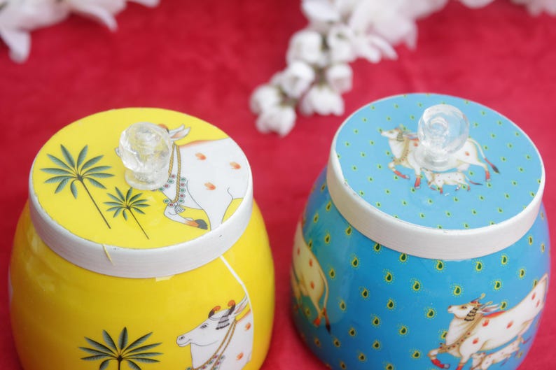 May include: Two decorative ceramic jars, one yellow and one blue, with white rims and clear glass knob handles. The yellow jar features palm tree and cow illustrations, while the blue jar has a cow and dot pattern. The jars sit on a red surface.