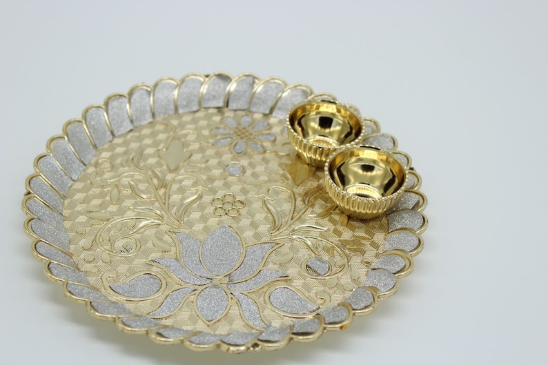 May include: A gold-coloured metal serving tray with a decorative floral design and two small gold-coloured bowls. The tray has a scalloped edge and a glittery finish.