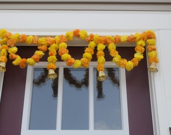 May include: A decorative door hanging garland featuring a series of vibrant yellow and orange artificial marigold flowers arranged in a draped pattern, accented with four metallic gold-toned conical bells hanging at intervals along the length.