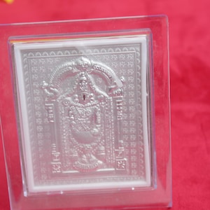 May include: A silver-colored, rectangular religious icon encased in a clear, square frame. The icon features a detailed embossed image of a deity, with intricate patterns and designs. The frame is set against a red background.