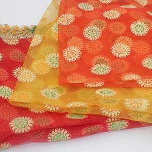 May include: Three colorful sheer scarves in red, orange, and yellow, each adorned with a repeating floral pattern. The edges are trimmed with gold and green decorative trim. The scarves are layered on top of each other.