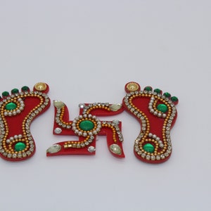 May include: Three red, decorative, wooden cutouts of a swastika and two footprints. The cutouts are decorated with gold and white beads and green gems.