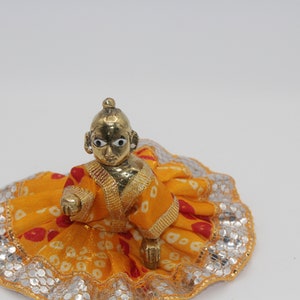 May include: A small golden figurine of a child wearing a yellow and red patterned dress with silver sequins. The figurine has large eyes and a serene expression.