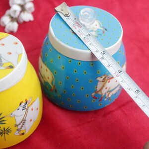 May include: Two decorative ceramic jars, one yellow and one blue, with lids. The jars feature a cow design and are being measured with a tape measure. The blue jar is approximately 4 inches in diameter.