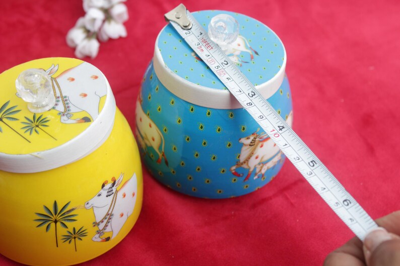 May include: Two decorative ceramic jars, one yellow and one blue, with lids. The jars feature a cow design and are being measured with a tape measure. The blue jar is approximately 4 inches in diameter.