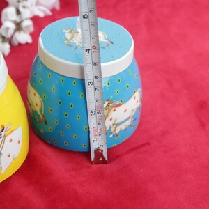 May include: Two decorative ceramic jars. One is yellow with a cow illustration, the other is blue with a cow illustration and measures approximately 3 inches (7.6 cm) in height. Both have white lids.