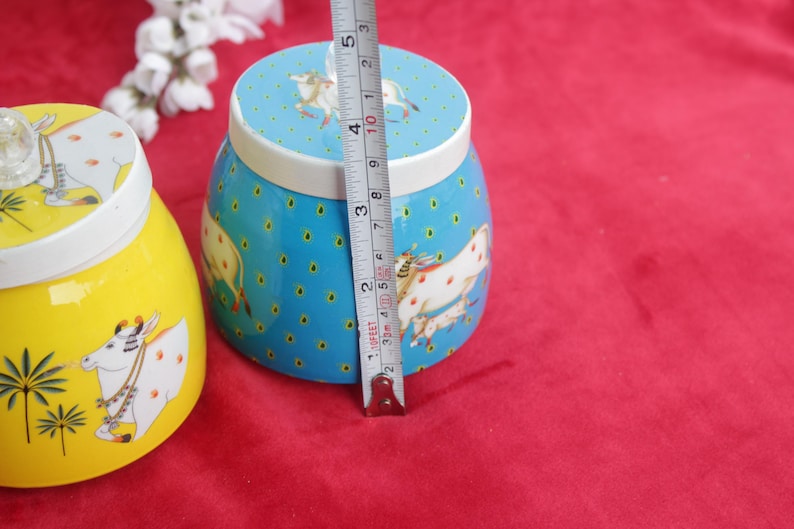 May include: Two decorative ceramic jars. One is yellow with a cow illustration, the other is blue with a cow illustration and measures approximately 3 inches (7.6 cm) in height. Both have white lids.