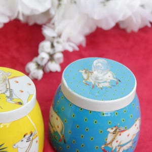 May include: Two decorative ceramic jars with lids. One jar is yellow with a cow and palm tree design, the other is blue with a cow design. Both have clear glass knobs on top. The jars sit on a red surface.