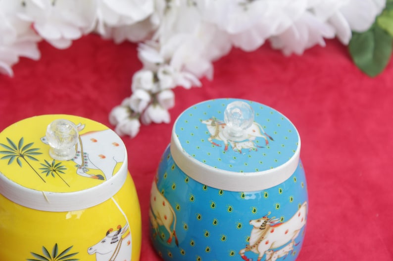 May include: Two decorative ceramic jars with lids. One jar is yellow with a cow and palm tree design, the other is blue with a cow design. Both have clear glass knobs on top. The jars sit on a red surface.