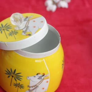 May include: A yellow ceramic jar with a lid, featuring a cow and palm tree design. The lid is off, revealing the inside of the jar. The jar has a clear knob on top. The background is red.