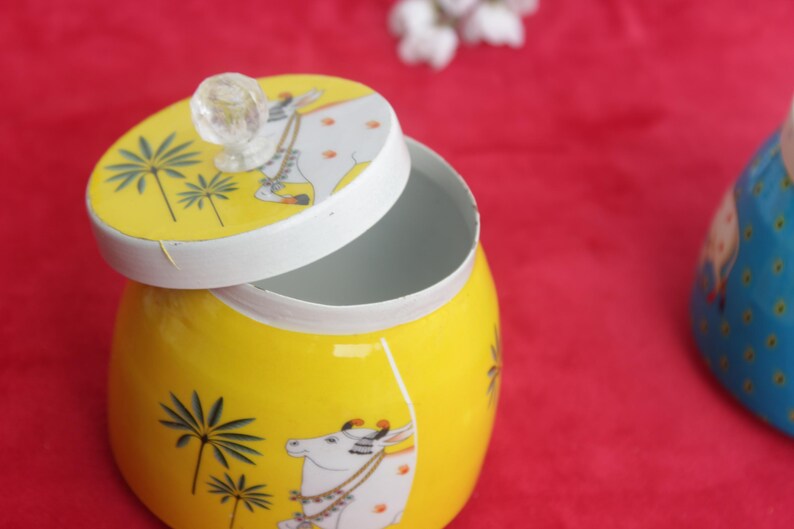 May include: A yellow ceramic jar with a lid, featuring a cow and palm tree design. The lid is off, revealing the inside of the jar. The jar has a clear knob on top. The background is red.