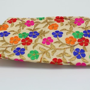 May include: A beige clutch with colorful embroidered flowers in red, pink, blue, orange, and green. The floral design is accented with gold-colored embroidery. The clutch has a gold cord handle and a tassel.