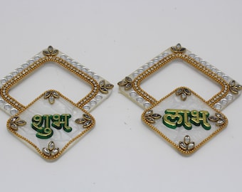 May include: Two white square-shaped decorative plaques with gold and silver embellishments. The top plaque has the word "Shubh" written in green and gold lettering. The bottom plaque has the word "Laxmi" written in gold lettering.