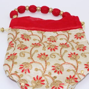 May include: A small, cream-colored drawstring bag with red and gold floral embroidery. The bag has a red fabric handle with gold beads.