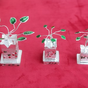 May include: Three silver-colored decorative plant sculptures with green and white leaves and stems. Each plant is in a clear, square-shaped pot with red and black accents, sitting on a white base. The background is a solid red color.