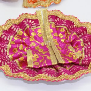 May include: A vibrant pink and gold decorative garment, likely a dress, with intricate gold floral patterns and trim. The edges are adorned with a gold lace-like border. The fabric has a shiny, satin-like appearance.