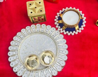 May include: A collection of decorative items on a red surface. Includes a silver-colored plate with two small gold bowls, a candle holder with a white candle, and a small gold box. A red fabric with gold patterns is in the background.