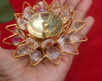 May include: A gold-coloured metal candle holder with a floral design, decorated with clear, faceted crystals. The candle holder has a central well for the candle and is held in a hand against a red background.