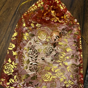 May include: A red and gold floral drawstring bag made of sheer fabric. The bag is decorated with a gold floral pattern and has a drawstring closure.