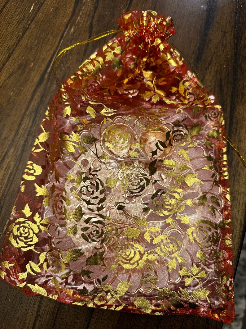 May include: A red and gold floral drawstring bag made of sheer fabric. The bag is decorated with a gold floral pattern and has a drawstring closure.