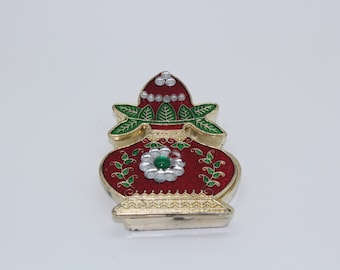 May include: A small, decorative, red and gold metal pot with green leaves and white pearls. The pot has a green and white flower design on the front.