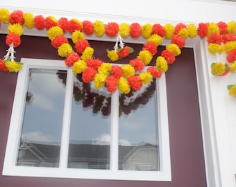 May include: A decorative door garland featuring a series of vibrant yellow and orange artificial marigold flowers arranged in a layered, draped pattern. The floral arrangement hangs across the top of a window frame with vertical hanging sections.
