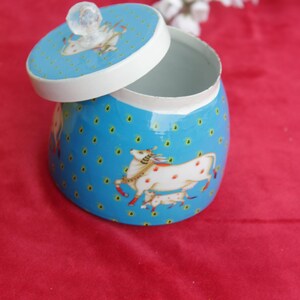 May include: A blue decorative jar with a lid, featuring a white rim and a clear crystal knob. The jar is adorned with a pattern of small yellow dots and illustrations of white horses with red spots. The lid is slightly ajar.