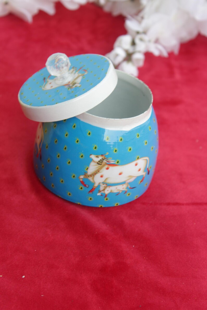 May include: A blue decorative jar with a lid, featuring a white rim and a clear crystal knob. The jar is adorned with a pattern of small yellow dots and illustrations of white horses with red spots. The lid is slightly ajar.