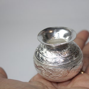 May include: A small, silver-colored, round pot with a wide, flared rim. The pot is held in a hand and features intricate floral and leaf patterns etched around its body. The pot has a shiny, reflective surface.