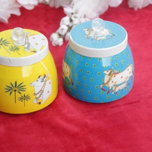 May include: Two ceramic jars with lids. One is yellow with a cow and palm tree design, the other is blue with a cow design. Both have white rims and clear knob handles. The jars sit on a red surface.